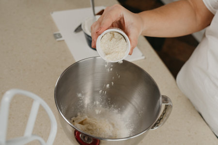 A close photo of the hands of a young woman who is putting an almond flour from the cup into the stainless steel bowl of the mixer.の写真素材