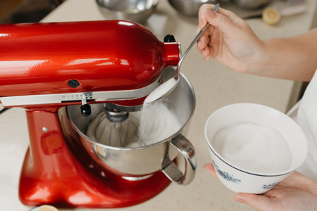 A close photo of the hands of a young woman who is spooning sugar to stainless steel soup bowl with the egg whites while the red stand mixer is whipping it.の写真素材