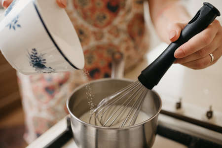 A close photo of a hand of a young woman who is mixing by the corolla cream, crushed zest, lemon juice and adding sugar into the saucepan on the gas stoveの写真素材