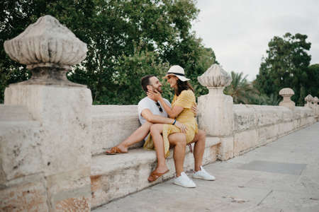 A girl in a hat and a yellow dress with a plunging neckline is sitting on the legs of her boyfriend with a beard on the ancient bridge in old Spain town. A couple of tourists on a date in Valencia.の写真素材