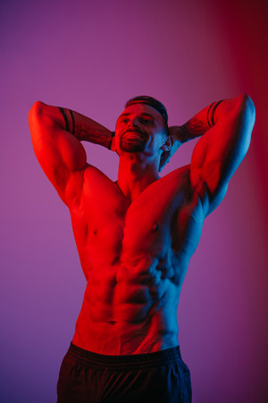 A muscular man with a beard is posing with arms behind his head under the blue and red light. An athletic guy is demonstrating his sporty torso. An athlete in shorts after a workout.の写真素材