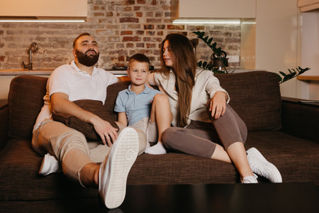 Dad with a beard, son, and young mom with long hair are watching TV and smiling on the sofa. The family is enjoying a happy evening at home. A child is listening to his mother.の写真素材