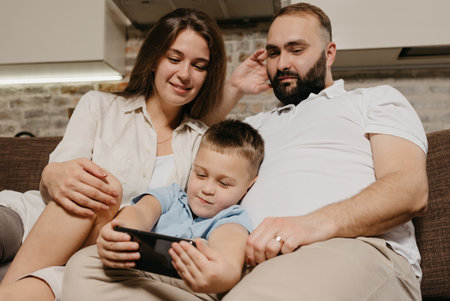 A dad, a son, and a mom are watching video on the sofa. A child is demonstrating a cartoon on the smartphone to mother and father in the evening at cozy home.の写真素材