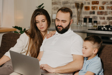 A father with a beard is talking about his achievements at work to a son and a wife at home. A family on the sofa in the evening. Dad is working remotely on a computer between relativesの写真素材