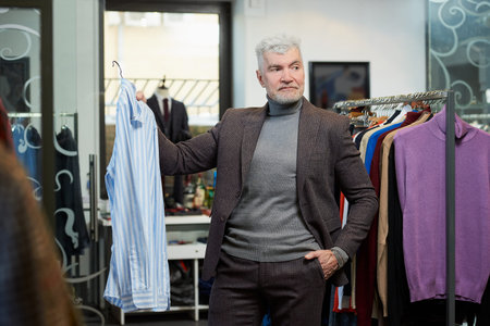 A mature man with gray hair is showing a shirt in a clothing store. A male customer with a beard wears a wool suit in a boutique.の写真素材
