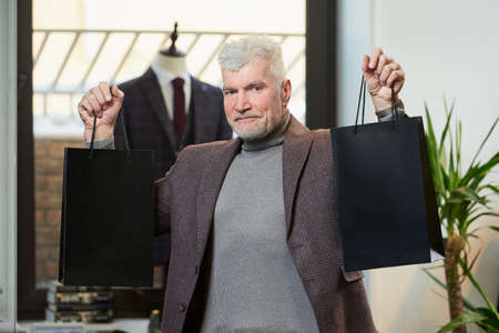 A happy mature man with gray hair and a sporty physique is showing two black paper bags with purchases in a clothing store. A male customer with a beard wears a wool suit in a boutiqueの写真素材