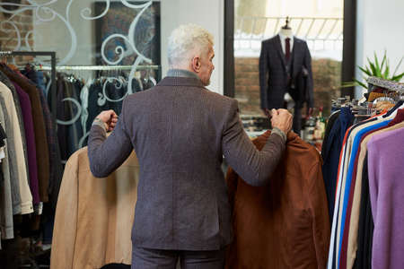 A photo from behind of a mature man with gray hair and a sporty physique is going holding suede jackets to the fitting room in a clothing store. A male customer with a beard wears a suit in a boutiqueの写真素材