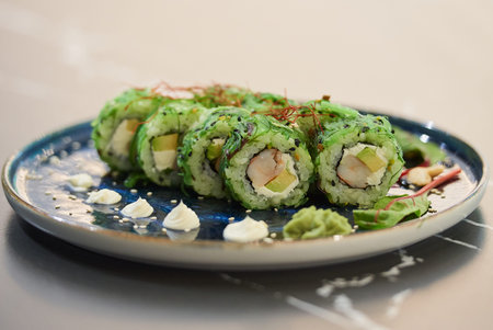 A close-up photo of sushi rolls with Chuka wakame seaweed, Philadelphia cheese, and shrimps on a blue ceramic plate with sauce, cashew, and wasabi on a black stone table.の写真素材