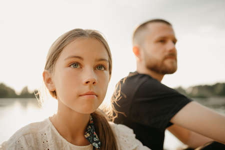 A close portrait of a young girl who is posing with her father with a beard on the coast of the lake. Single-parent stylish family on vacation on the sunset.の写真素材