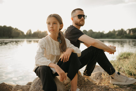 A young girl is posing on stones with her father with a beard and sunglasses on the coast of the lake. Single-parent stylish family on vacation on the sunset.の写真素材