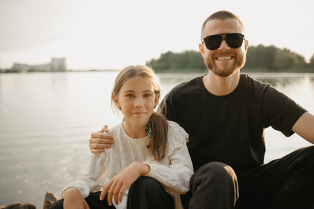 A close portrait of a young smiling girl who is posing with her father with a beard who is hugging her on the coast of the lake. Single-parent family on vacation on the sunset.の写真素材