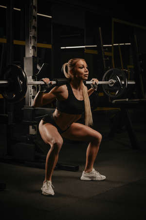 A fit woman with blonde hair is squatting with a barbell near the squat rack in a gym. A girl is doing a leg workout.の写真素材