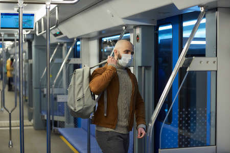 A man with a beard in a face mask to avoid the spread of is putting on a gray backpack while riding a subway car. A bald guy in a surgical mask is keeping social distance on a train.の写真素材