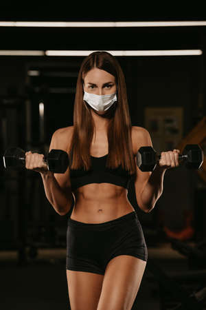 A fit woman in a face mask to avoid the spread of coronavirus is doing bicep curls with dumbbells. A sporty girl in a surgical mask is posing during the arms workout in a gym.の写真素材