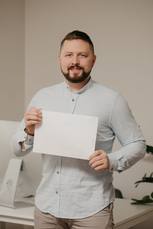 A smiling man with a beard is standing with an empty sheet of white paper near a steel desktop...