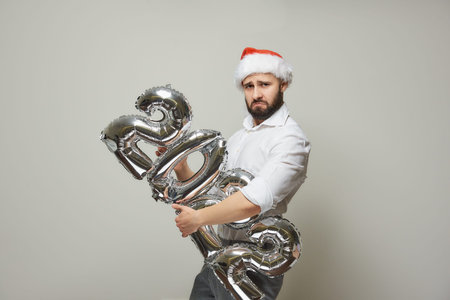A proud man in a red velvet Santa hat is holding silver balloons in the shape of 2022. A happy guy...