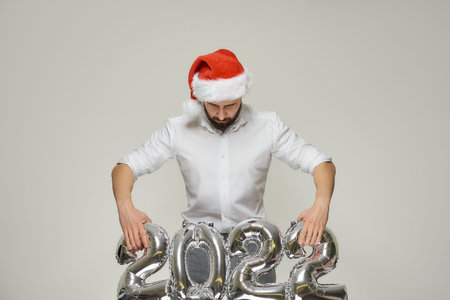 A man with his head down in a red velvet Santa hat is lifting silver balloons in the shape of 2022....