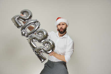 A dashing man in a red velvet Santa hat with his lips in a tube is pushing silver balloons in the shape of 2022 to the vertical left. A guy with a beard at a New year party.の写真素材