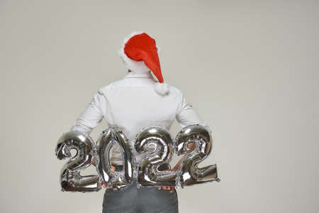 A man in a red velvet Santa hat is lifting behind his back silver balloons in the shape of 2022. A guy with a beard at a New year party.の写真素材