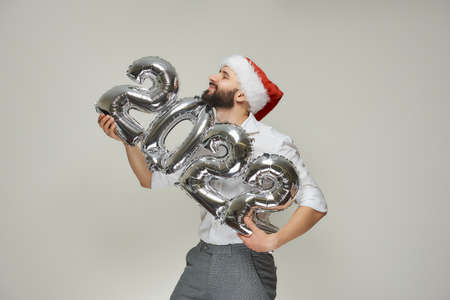 A smiling man in a red velvet Santa hat is hugging silver balloons in the shape of 2022 gently. A happy guy with a beard at a New year party.の写真素材