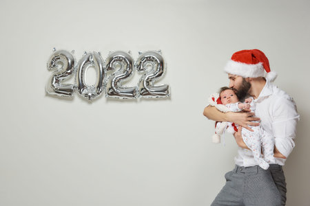 A happy father in a red velvet Santa hat is holding his infant daughter near silver balloons in the...