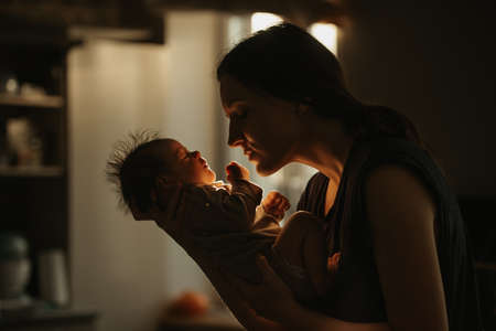 A medium long shot of the mother who is holding the newborn baby in her hands and hugging her at home. The infant and the mom in the sunlight in front of the window in the evening.の写真素材