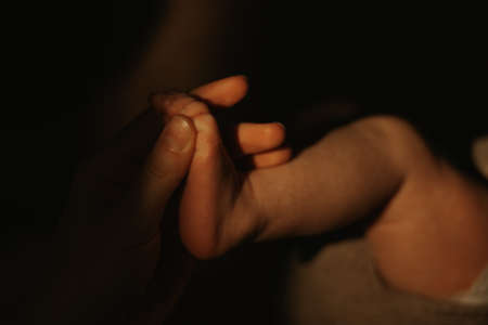 A close-up photo of the hand of the mother who is massaging the foot of a newborn baby. An infant in a bodysuit is lying on a sofa in the sunlight in the evening.の写真素材