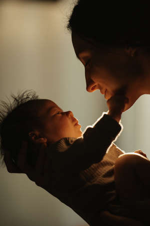 A close up shot of the happy mother who is holding the newborn baby in her hands and hugging her. The smiling infant is reaching out for the mom in the sunlight in front of the window in the evening.の写真素材