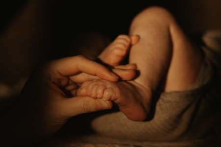 A close-up photo of the hand of the mother who is caressing the foot of a newborn baby. An infant in a bodysuit is lying on a sofa in the sunlight in the evening.の写真素材