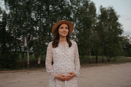 A medium long shot of a pregnant woman who is holding her belly on the sand road. A happy pregnancy on the farm on the sunset. A lady in a long dress and a hat is in the park in the evening.の写真素材