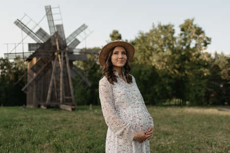 A medium long shot of a pregnant woman in the windmill background who is holding her belly with her hands on the farm. A happy pregnancy. A lady in a long dress and a hat at the meadow in the evening.の写真素材