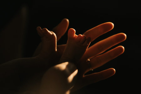 A close-up photo of the gentle hand of the mother who is holding the feet of her newborn baby. The...