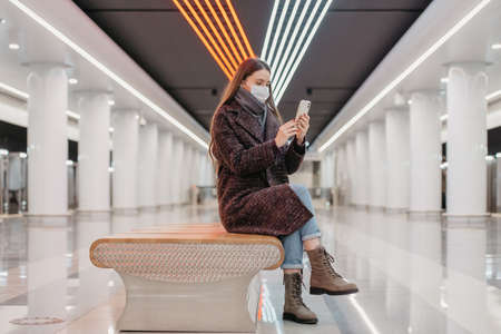 A woman in a medical face mask is sitting in the center of the subway platform with a smartphone and doing a selfie. A girl with long hair in a surgical mask is keeping social distance in the metro.の写真素材