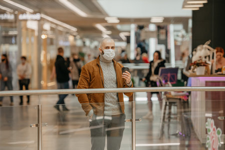 A man in a face mask to avoid the spread of coronavirus is holding a cup of coffee in the shopping center. A bald guy in a surgical mask is keeping social distance.の写真素材