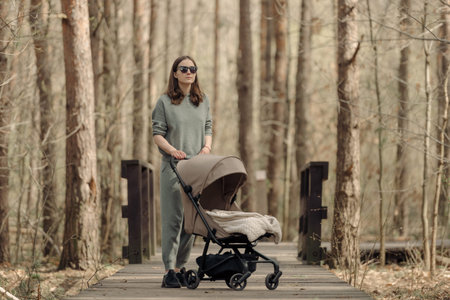 The young mom is walking on the wooden walkway with her infant child who is sleeping in the baby...