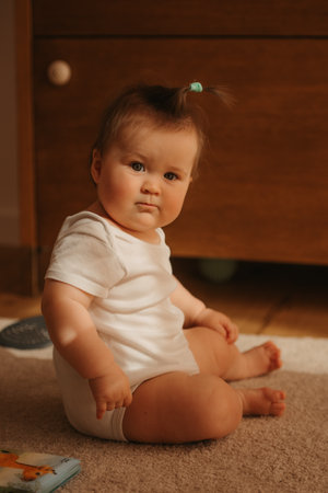 A 7-month girl is sitting near a dresser in a bodysuit at home.の写真素材