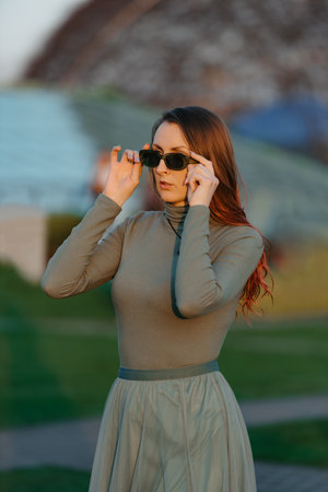 A redhead woman in green clothes is checking her sunglasses in the park at sunset.の写真素材
