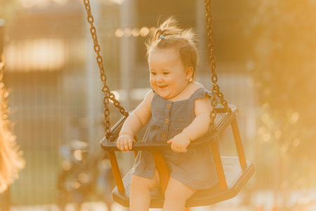 A happy toddler baby girl in a dress on a swing on the warm summer eveningの写真素材