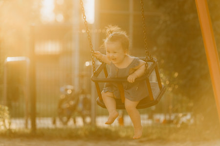 A happy toddler baby girl in a dress on a swing on the warm summer eveningの写真素材