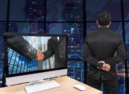 the computer on the wood table with Hand shake between businessman at the screen in front of the glass window over on the trading graph over the blurred photo of cityscape,Back side of Businessmanの写真素材