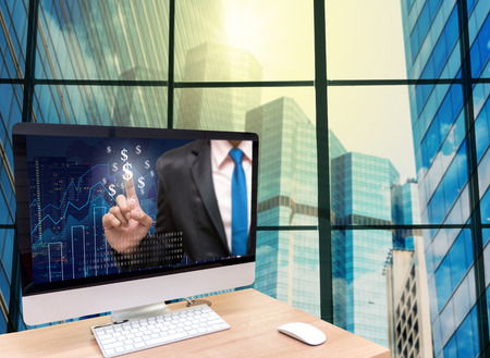 the computer on the wood table with Businessman point sign of money on the trading graph at the screen in front of the glass window over the blurred photo of cityscape backgroundの写真素材