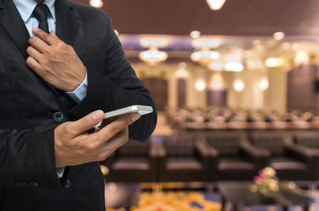 Businessman using the smart phone on the Abstract blurred photo of conference hall or seminar room backgroundの写真素材