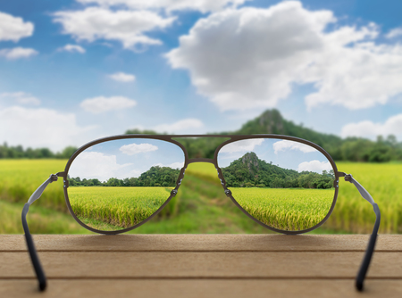 Conceptual image of Landscape focused in glasses lenses over the photo blurred of Rice field with mountain background under blue sky with cloudの写真素材