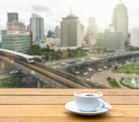 White coffee cup on wood table beside the window which can see the abstract Blurred photo of sky train with cityscape background, business traffic in rush hour conceptの写真素材