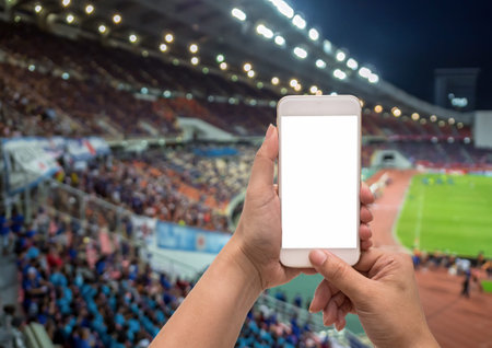 Female hand holding mobile smart phone touch screen on Abstract blurred photo crowd of spectators on a stadium with a football match, sport background conceptの写真素材