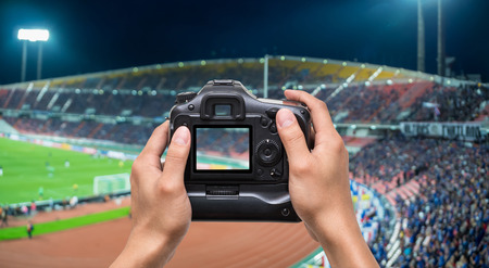 Hand holding the camera over Abstract blurred photo crowd of spectators on a stadium with a football match, sport background conceptの写真素材