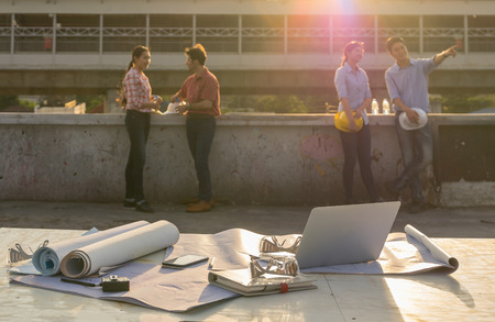 Construction equipment such as blueprint, Safety glasses and technology laptop over the photo blurred of engineers working with sun ray and lens flare background, industrial concept, worm color toneの写真素材