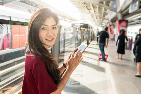 Asian woman passenger with casual suit using the technology tablet with lens flare in the BTS Skytrain rails or MRT subway for travel in the big city, lifestyle and transportation conceptの写真素材