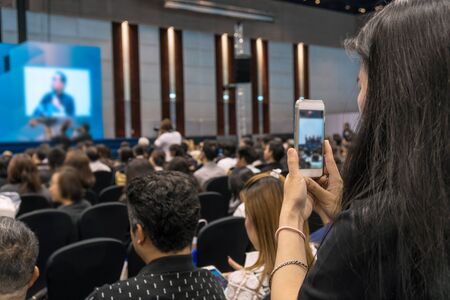 Speakers on the stage with Rear view of Audience taking photo or video for Live in the conference hall or seminar meeting, business and education about investment concept