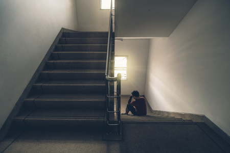 Depressed man sitting head in hands on the staircase in the fire escape or building stair with low light environment, dramatic scene conceptの写真素材